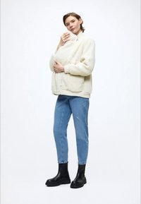 Giacca in ecopelliccia color crema con collo alto e tasche frontali, abbinata a jeans azzurri e stivaletti neri. Design semplice e tessuto dalla texture.