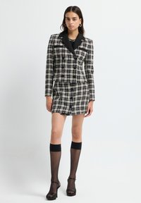 Abito blazer in tweed a scacchi bianco e nero con revers neri, due bottoni e un'orlo corto, abbinato a calze nere trasparenti fino al ginocchio e tacchi.