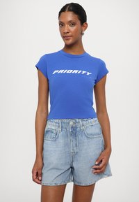 T-shirt di cotone blu con maniche corte e testo bianco "PRIORITY", abbinato a pantaloni corti di jeans blu chiaro con vita e tasche.