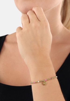 Bracciale in tono oro con piccole perle rosa, bianche e dorate, e un ciondolo a forma di cuore inciso con la parola "amor". Texture liscia, design delicato.