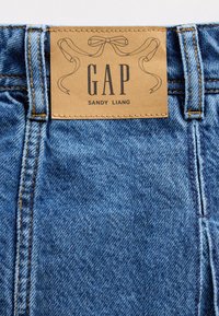 Lichtblauwe spijkerbroek met een leren label met de tekst "GAP SANDY LIANG" en decoratieve stiksels langs de taille en het ritsgebied.