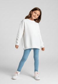 Biały, oversize'owy sweter, jasnoniebieskie legginsy i białe trampki z różowymi sznurówkami. Modelka stoi z jedną nogą do przodu, uśmiechając się. Proste tło.