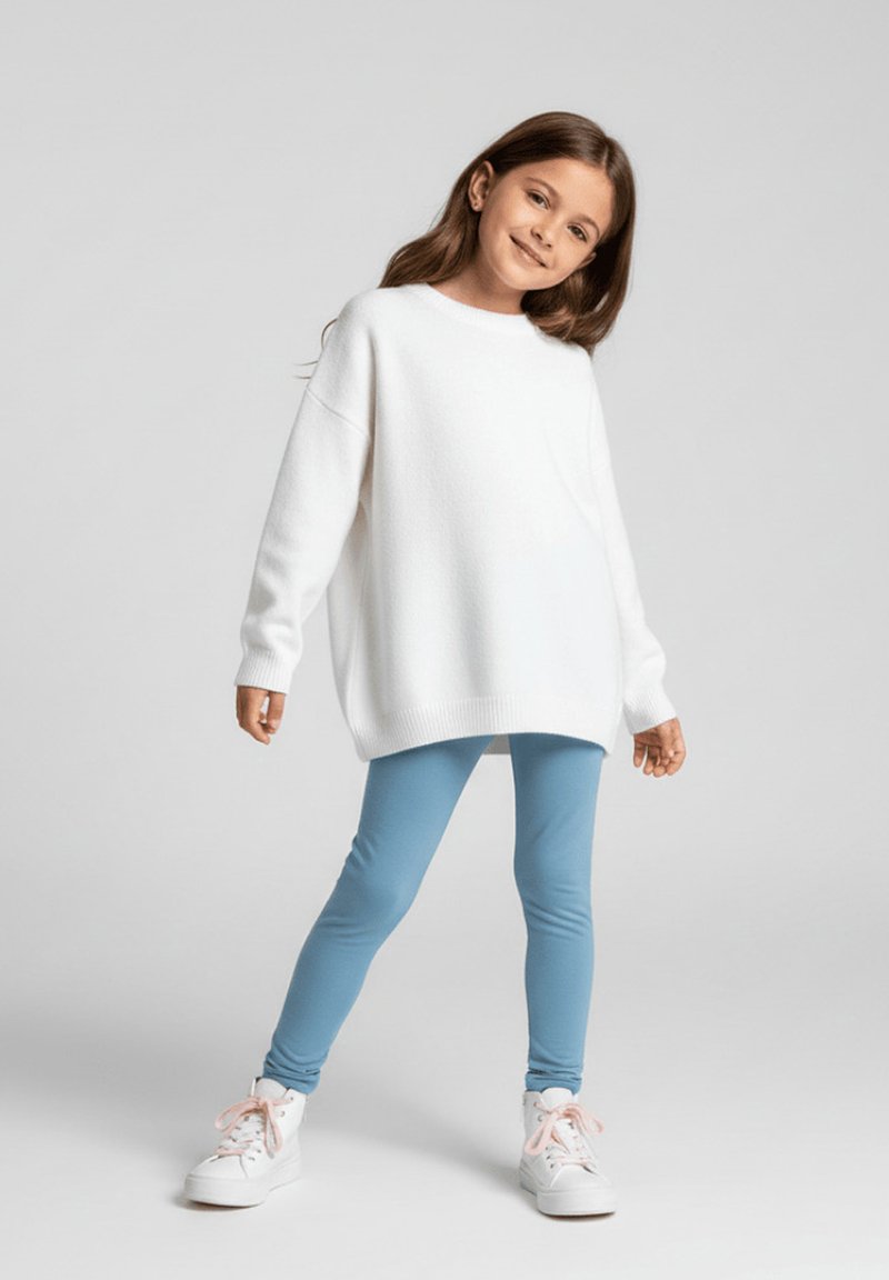 Biały, oversize'owy sweter, jasnoniebieskie legginsy i białe trampki z różowymi sznurówkami. Modelka stoi z jedną nogą do przodu, uśmiechając się. Proste tło.