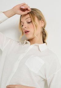 Camisa de lino blanca con cuello, cierre de botones y un solo bolsillo en el pecho. Presenta una textura ligera y un ajuste relajado.