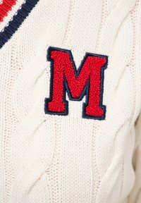 Biały sweter dzianinowy z wzorem w warkocze, z dużym czerwonym emblematem "M" obramowanym granatowym kolorem. Miękka tekstura z wyraźnym ściągaczem.