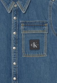 Koszula dżinsowa z klasycznym kołnierzykiem, srebrnymi guzikami zatrzaskowymi i kieszenią na piersi z czarną naszywką Calvin Klein Jeans.