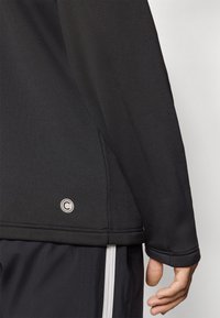 Camisa deportiva de manga larga negra con una tela texturizada. Presenta un logo circular cerca del dobladillo y costuras reforzadas para mayor durabilidad.