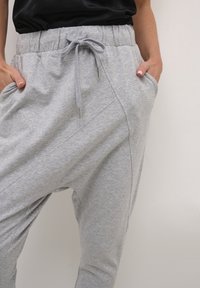 Persoon in lichtgrijze trainingsbroek met een strik in het koord en handen in de voorzakken tegen een effen achtergrond.