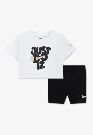 Biela skrátená tričko značky Nike s nápisom „Just Do It“ a béžovým logom Swoosh, kombinovaná s čiernymi textúrovanými šortkami s béžovým logom Nike Swoosh.