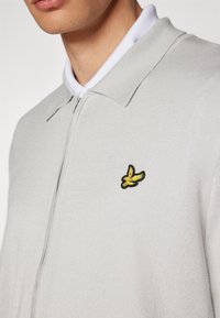 Gray knit zip-up sweater with a collared neckline, featuring a black and yellow embroidered logo of a bird on the left side.