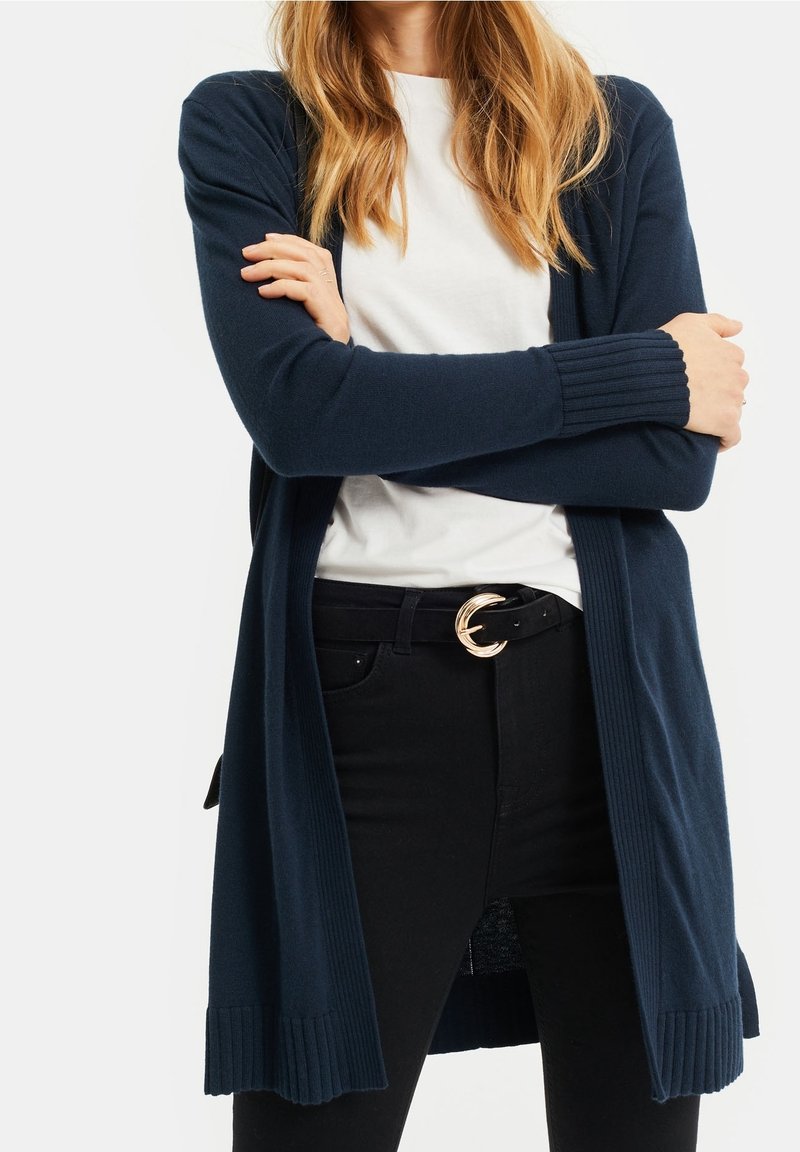 Marinblå lång cardigan i mjukt stickat tyg med ribbade ärmslut och nederkant, bärs över en vit t-shirt och svarta jeans. Guldspänne på bältet som accent.