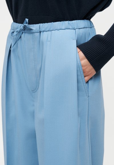 Personne portant un pantalon plissé bleu clair avec une taille élastique et un cordon de serrage, la main partiellement dans la poche, associée à une manche de pull côtelée noire.
