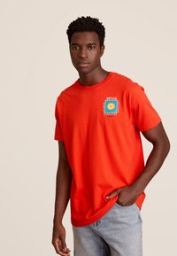 Rode katoenen T-shirt met een vierkante grafiek en een blauw zonnestraalontwerp, met de tekst "SICILIA" op de linker borst. Korte mouwen, ronde halslijn.