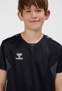 Ung pojke med kort brunt hår som ler, klädd i en svart hummel sporttröja med V-ringning och logotyp på bröstet, mot en enfärgad bakgrund.