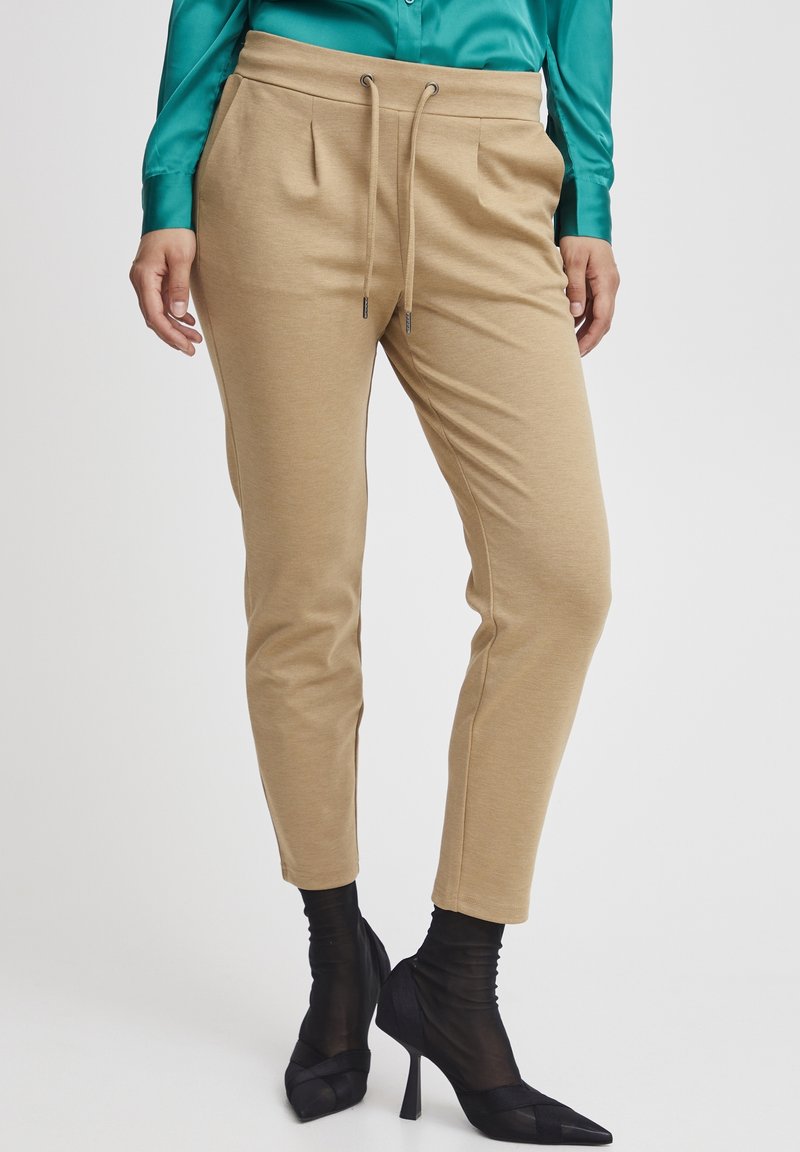 Femme portant un pantalon beige à cordon, un chemisier en satin bleu sarcelle, des chaussettes noires transparentes et des chaussures à talons hauts noires à bout pointu, debout.