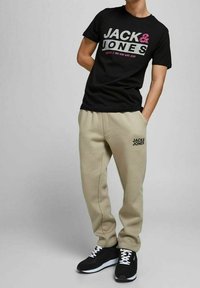 Černé tričko s grafikou "JACK & JONES" v šedé/růžové barvě, zkombinované s béžovými tepláky a černými sportovními botami s bílými tkaničkami.