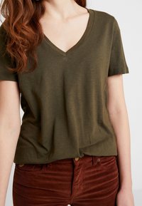 Olivově zelené tričko s výstřihem do V z měkké látky, v kombinaci s kalhotami z cordu v barvě rezavé, které mají vertikální pruhy a knoflíkové detaily.