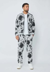 Modèle masculin portant une veste à motif floral noir et blanc et un pantalon assorti, un t-shirt blanc et des baskets blanches, se tenant devant un fond uni.