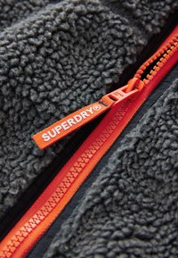 Close-up of a gray fleece jacket with an orange zipper and a pull tab branded with "SUPERDRY®" in white text.