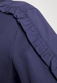 Námořnická modrá blůza s volánkovým detailem na rameni. Látka vypadá hladká s mírným leskem. Volán přidává texturu a vizuální zajímavost.