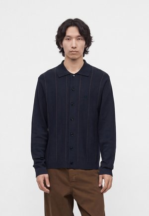 Ung mand med mørkt mellem-langt hår iført en marineblå cardigan med knapper med subtile lodrette striber og brune bukser, stående mod en hvid baggrund.