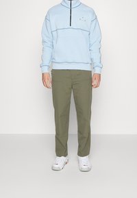 Světle modrá fleecová mikina s polovicí zipu a přední kapsou, sladěná s olivově zelenými kalhotami a bílými botami s červenými akcenty.