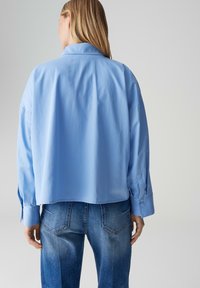 Camicia corta blu con una texture morbida, caratterizzata da maniche lunghe e colletto classico. Abbinata a jeans blu scoloriti, con distintive tasche posteriori.