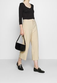 Svart långärmad topp, beige vida byxor och svarta loafers. Håller en liten svart väska med texturerad yta. Vit bakgrund.