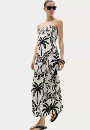 Kvinde iført sort-hvid stropløs maxikjole med palme- og blomsterprint, sorte sandaler, solbriller og armbånd.