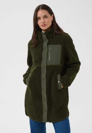 Femme portant une longue veste polaire vert foncé avec des boutons-pression, une poche poitrine zippée et les mains dans les poches latérales.