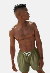 Zöld úszónadrág fehér zsinórral, sima anyagból készült. A modell póló nélkül van, bemutatva egy tetoválást a mellkasán.