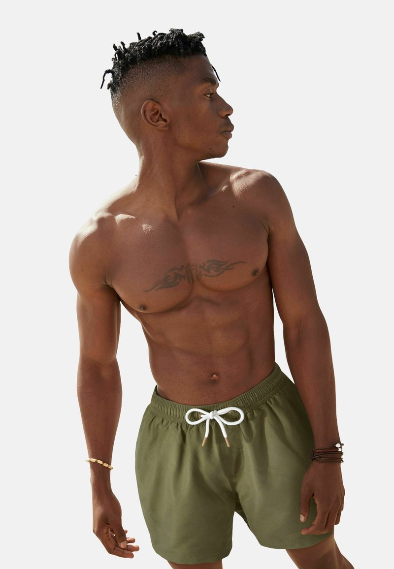 Zöld úszónadrág fehér zsinórral, sima anyagból készült. A modell póló nélkül van, bemutatva egy tetoválást a mellkasán.