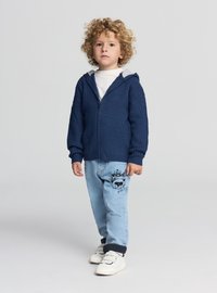 Felpa con cappuccio e zip color blu navy in tessuto strutturato, abbinata a jeans azzurri con un motivo di orso sulla gamba sinistra e risvolti arrotolati.