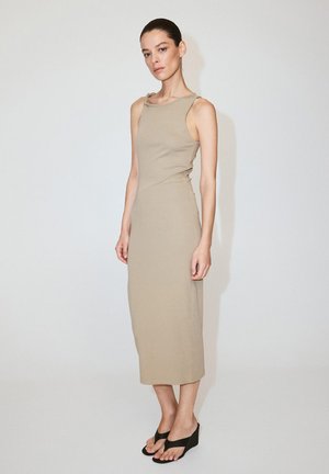 Femme aux cheveux tirés en arrière, portant une robe midi beige sans manches et des sandales compensées noires, debout contre un mur clair uni.