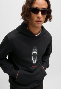 Svart hettegenser med grafikk av en brølende munn, med hvite tenner. "HUGO" er trykt i rødt under designet. Glatt stoff, kengurulomme.