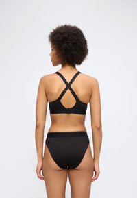 Γυναίκα με κυματιστά μαλλιά, φοράει μαύρο μαγιό με ψηλόμεσο κάτω μέρος και μαύρο σουτιέν με λουράκια που διασταυρώνονται στην πλάτη, στέκεται μπροστά σε λευκό φόντο.