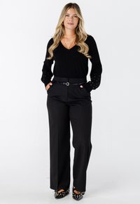 Zwarte lange mouwtop met versierde manchetten, gecombineerd met zwarte gestreepte wijde broek en zwarte gespijkerde hakken, wat een monochromatische look creëert.