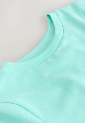 Artimųjų planas žalios mėtos spalvos medvilninio marškinėlio apykaklės ir peties ant šviesiai pilkos spalvos fono.