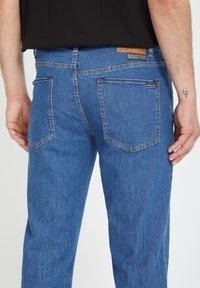 Jean azul de denim con corte recto, que cuenta con bolsillos traseros, costuras en contraste y una etiqueta de cuero con marca en la cintura.