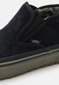 Schwarzer Slipper aus Wildleder mit elastischen Seitenpaneelen und grünem Gummisohlen. Das Innenfutter zeigt ein grün-schwarzes Muster. Vans-Logo ist vorhanden.