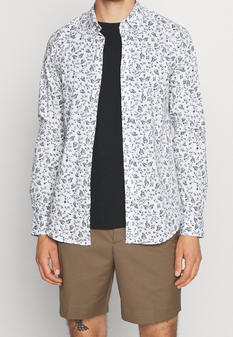 Chemise blanche à boutons avec un petit motif floral noir, portée sur un t-shirt noir. Associée à un short marron. Tissu lisse, design décontracté.