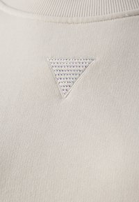 Kermaansävyinen neulottu kangas, jossa on resoripinta. Siinä on pieni kolmionmuotoinen embleemi, joka on koristeltu monivärisillä strassikivillä.