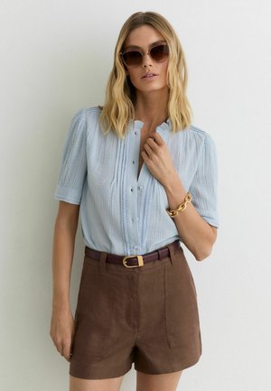 Femme aux cheveux blonds portant des lunettes de soleil, un chemisier bleu clair boutonné, un short marron avec ceinture et un gros bracelet en or, debout contre un mur blanc.