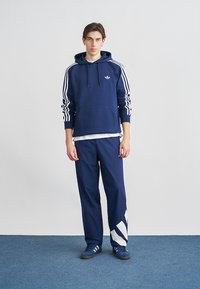 Jonge man die een marineblauwe Adidas hoodie draagt, bijpassende trainingsbroek met witte strepen, en blauwe sneakers op een blauw tapijt.