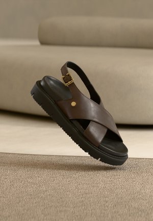 Smeđa kožna sandala otvorenih prstiju s ukriženim trakama, zlatnom kopčom i debelim crnim potplatom, postavljena iznad teksturiranog tepiha i pozadine bež kauča.