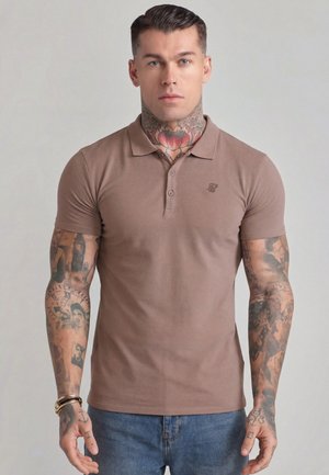 Nyakán és karjain tetoválásokkal rendelkező férfi, barna galléros pólót és kék farmert viselve, egy egyszínű világos háttér előtt áll.