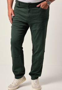 Uomo che indossa pantaloni jogger verde scuro con polsini elasticizzati e sneakers bianche, mano in tasca e un orologio al polso sinistro.