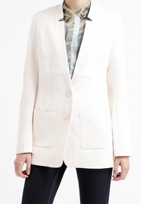 White tailored blazer with two front pockets and two buttons, worn over a camouflage-patterned shirt. Smooth texture, structured design.