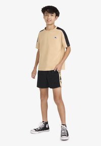 Chemise beige à manches courtes avec des accents noirs, portée avec un short noir présentant un marquage latéral jaune et des baskets montantes noires.