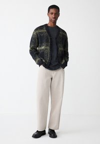 Pleteni cardigan v črno-zeleni karo vzorec z gumbi, kombiniran s temno sivo majico in svetlo bež širokimi hlačami ter črnimi čevlji.